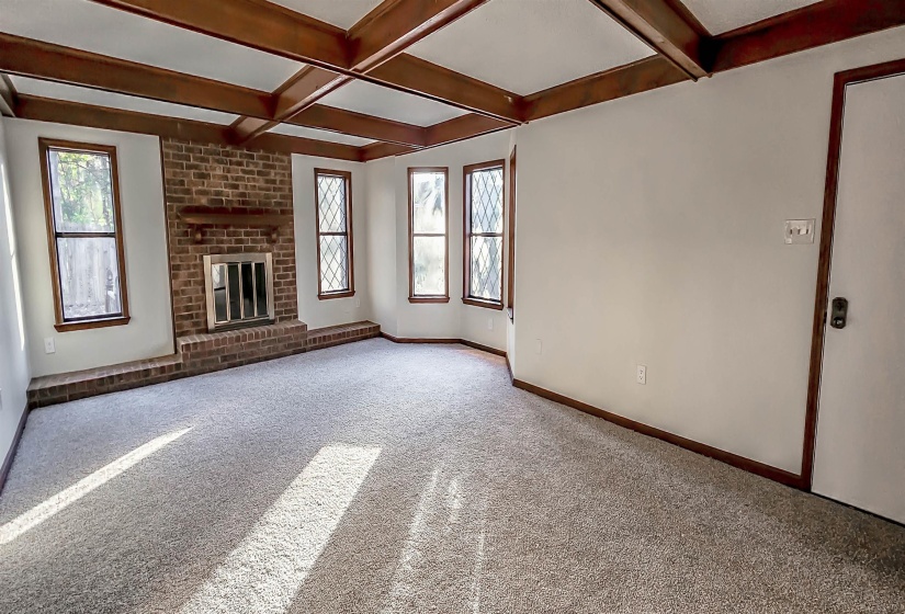 Spacious living area featuring a brick fireplace with a raised hearth, wood-beamed coffered ceiling, multiple windows, and light-toned carpeting