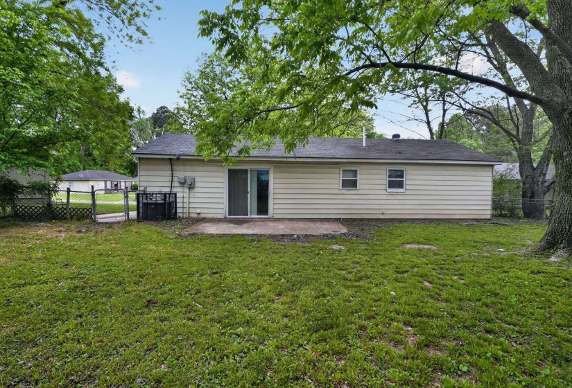 Expansive backyard featuring a concrete patio, sliding glass door, mature trees, and partial chain-link fencing