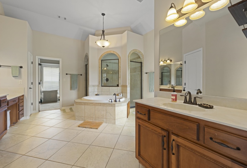 Spacious bathroom featuring a built-in soaking tub with tiled surround, a separate glass-enclosed shower, dual vanity with wood cabinetry and light-toned countertops, and large format tile flooring