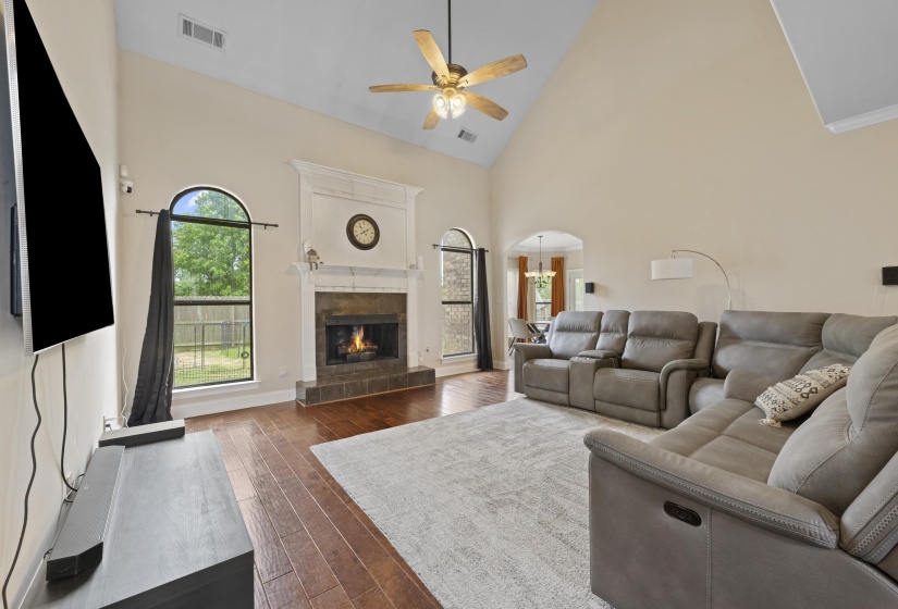 Living area featuring vaulted ceilings, wood-finish flooring, a stone-surround fireplace with a white mantel, arched windows, and a ceiling fan