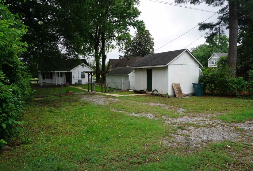 White siding structure with an attached shed, dark shingle roofing, and established perimeter trees