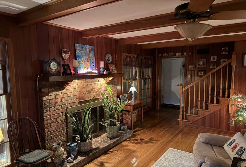 Brick fireplace with a wood mantel, wood-paneled walls, exposed wood beams, wood-finish flooring, and a wood staircase with balusters