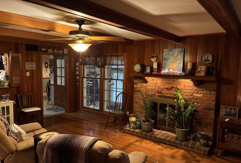 Brick-faced fireplace with a wood mantel, paneled walls, wood-finish flooring, exposed ceiling beams, and a ceiling fan with integrated lighting