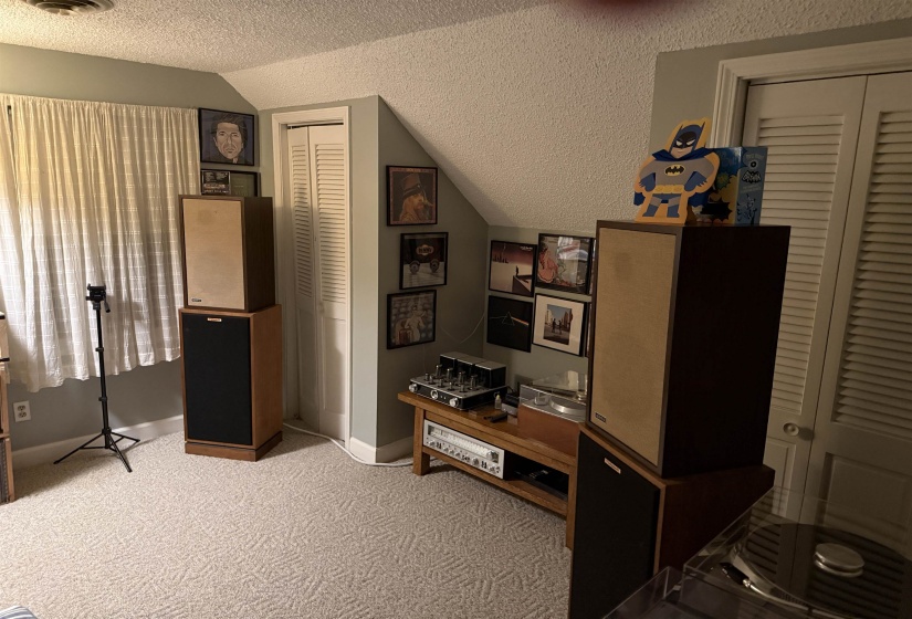 Carpeted interior space featuring a window with white curtains, a textured ceiling, a louvered bi-fold door, and an angled wall