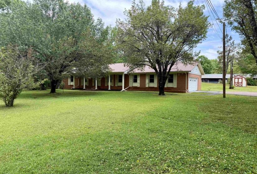 Brick-clad ranch home with a metal roof, featuring a covered porch with white columns and shutters, an attached garage, and a spacious front lawn with mature trees