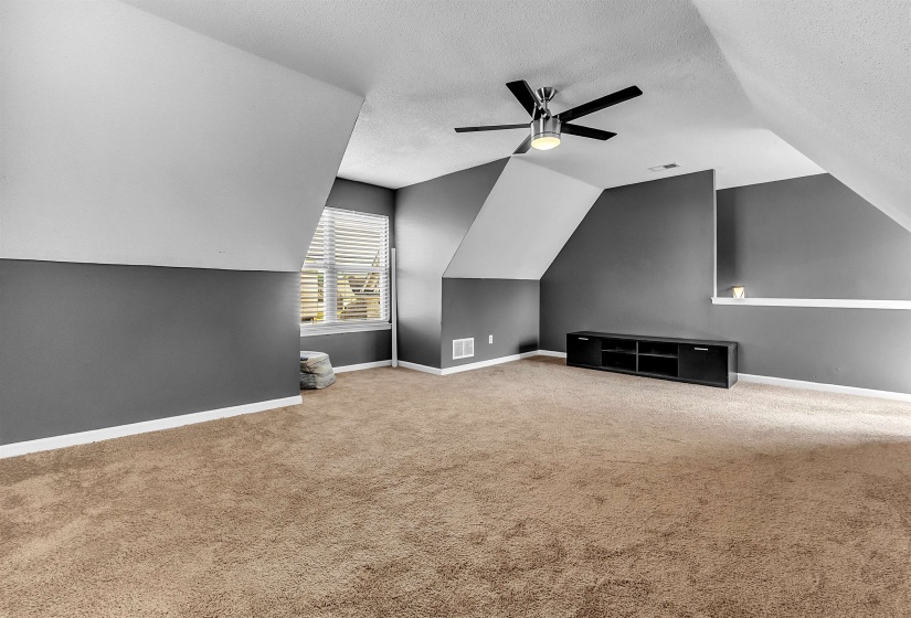Spacious room featuring sloped ceilings, plush carpet flooring, a window with horizontal blinds, and a contemporary ceiling fan