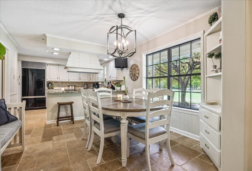 Open-concept kitchen and dining area featuring stone-finish floor tiles, a window with exterior visibility, white cabinetry, a built-in kitchen range hood, and recessed lighting