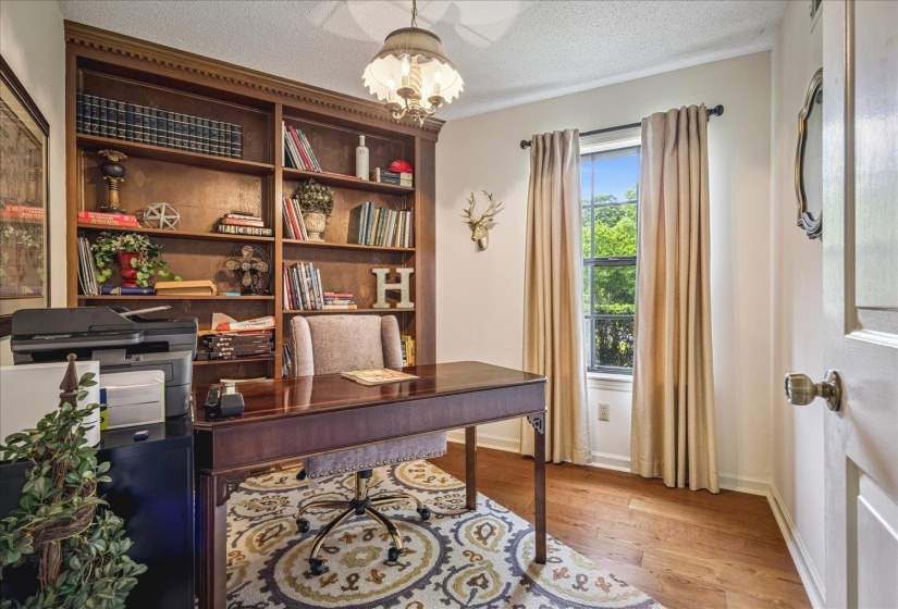 Dedicated workspace featuring extensive built-in wooden shelving, wood-finish flooring, a window with outdoor tree scenery, and a ceiling-mounted light fixture