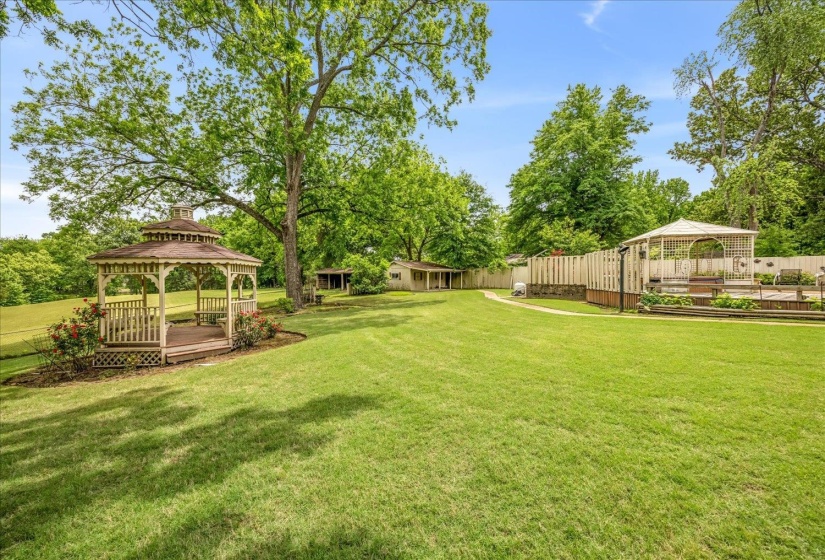 Expansive green lawn featuring a wooden gazebo with a shingled roof, a secondary covered patio area, mature trees, and landscaping elements