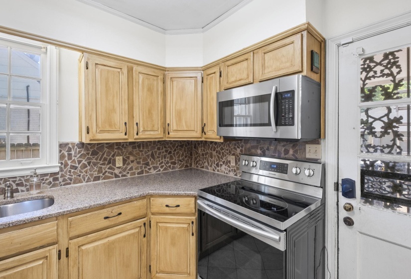 Kitchen area featuring light wood cabinetry, speckled countertops, a mosaic tile backsplash, stainless steel microwave, and a stainless steel electric range