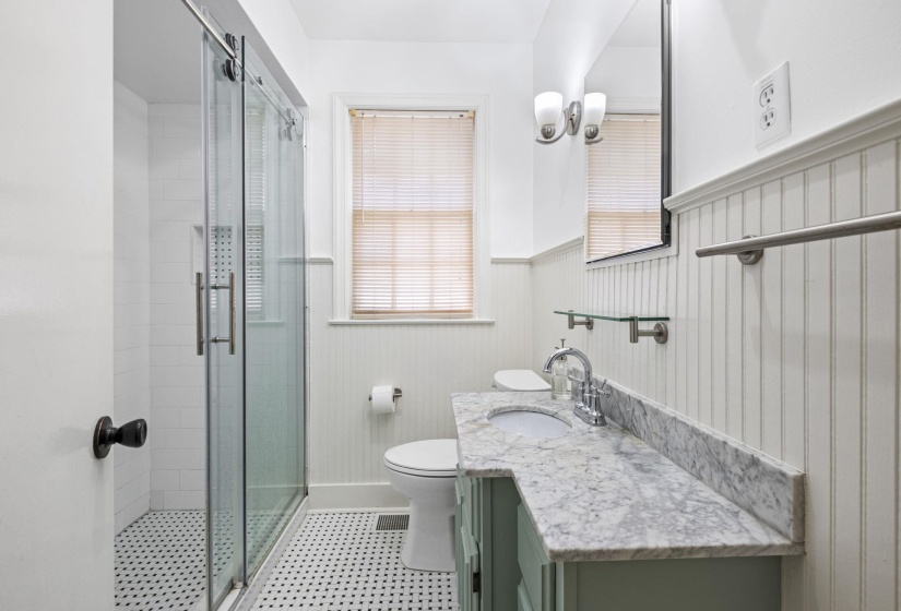 Bathroom featuring a glass-enclosed shower with subway tile, black and white mosaic floor tile, a vanity with a stone countertop, beadboard wainscoting, and a window with blinds