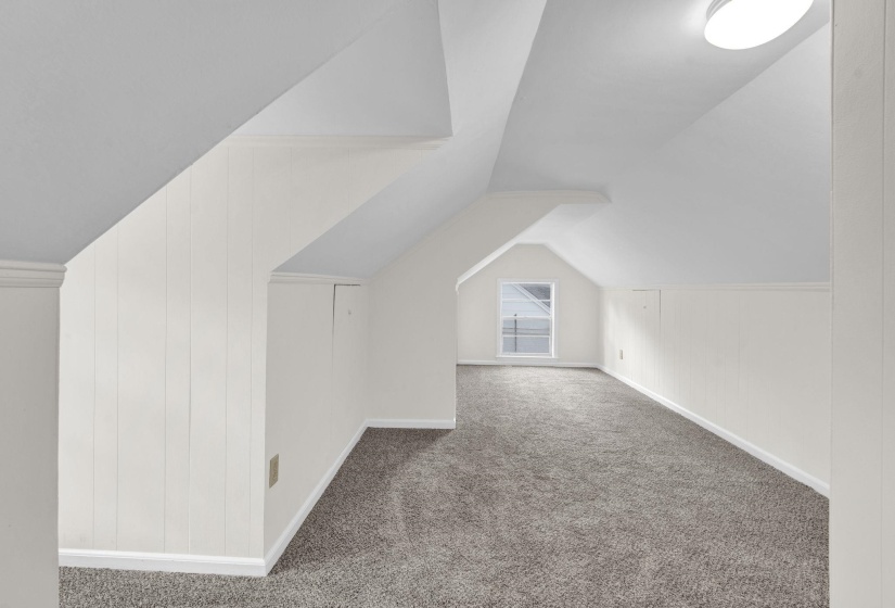Carpeted attic space featuring white paneled walls, a single window, and a flush-mount ceiling light