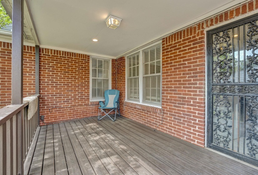 Expansive covered porch featuring a brick exterior wall, dark wood-finish flooring, white double-hung windows with blinds, and an ornate wrought iron security door