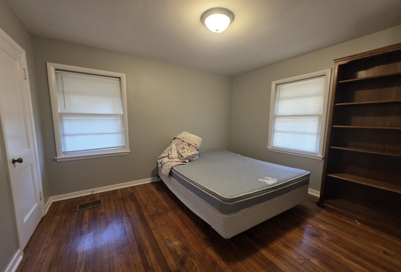 Bright room featuring two windows with horizontal blinds, wood-finish flooring, a flush-mount ceiling light fixture, and a built-in wood-finish bookshelf
