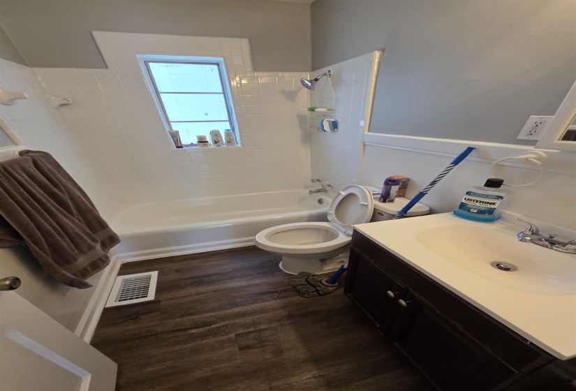 Bathroom featuring a white tub, shower with white subway tile surround, wood-finish flooring, dark wood vanity with integrated sink, and a white toilet