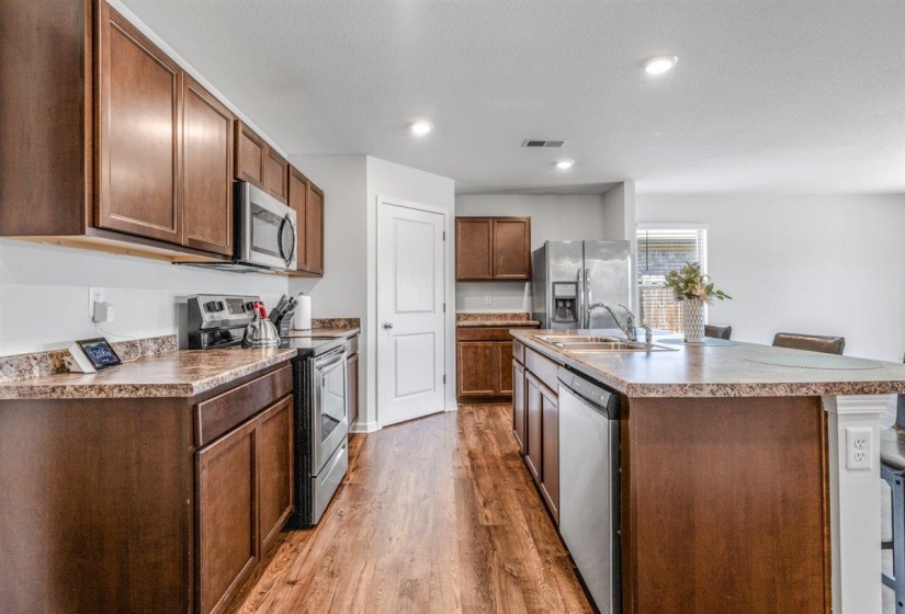 Kitchen featuring wood-finish flooring, dark wood cabinetry, light-toned countertops, stainless steel appliances, and a central island with an integrated sink
