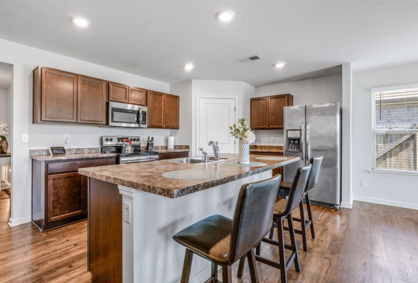 Bright kitchen featuring a breakfast bar island with an undermount sink, stainless steel appliances, wood-finish flooring, recessed lighting, and rich wood cabinetry