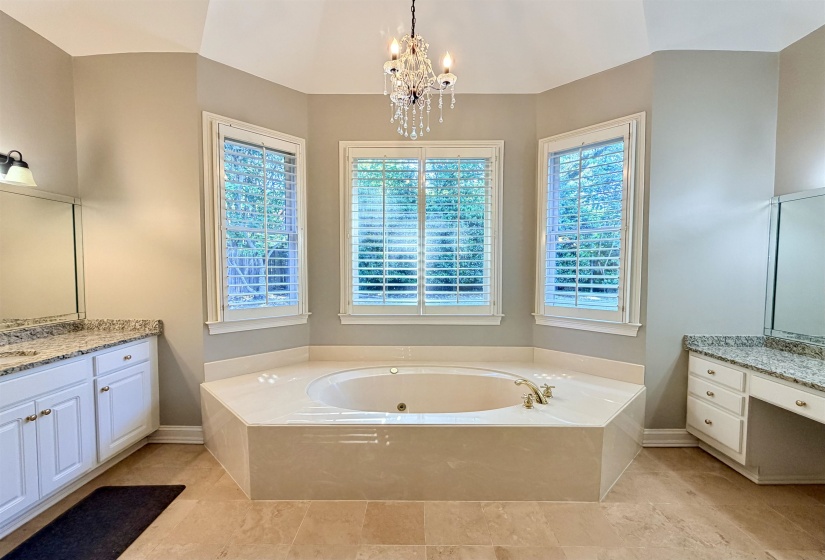 Spacious bathroom featuring a built-in soaking tub, plantation shutters, twin vanity areas with stone countertops, and a decorative chandelier