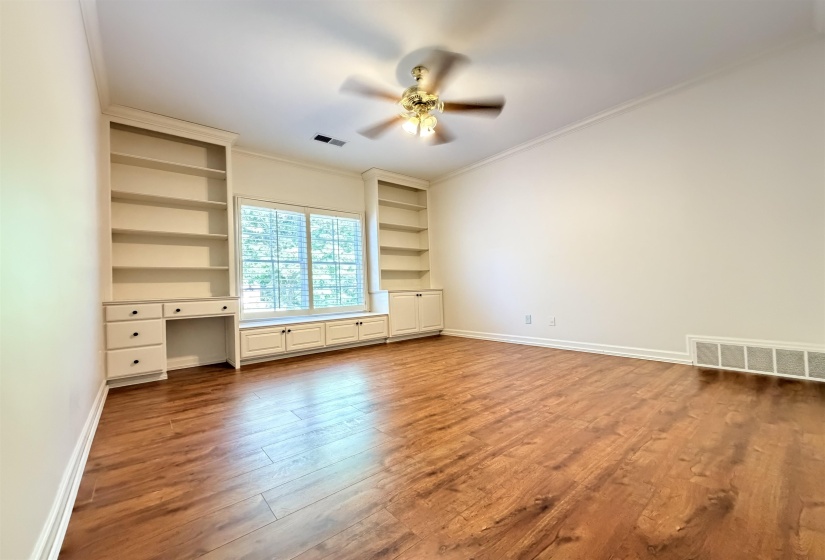 Spacious room featuring wood-finish flooring, a built-in desk with shelving, a window with a window seat, and crown molding