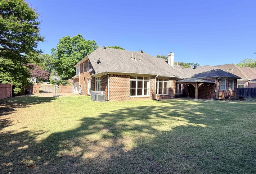 Spacious backyard featuring a large brick exterior, multiple windows, a covered patio structure, and a mature tree line