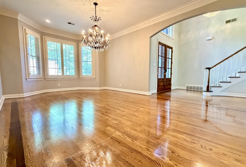 Hardwood flooring throughout a spacious dining room featuring a crystal chandelier, bay windows with plantation shutters, an arched entryway