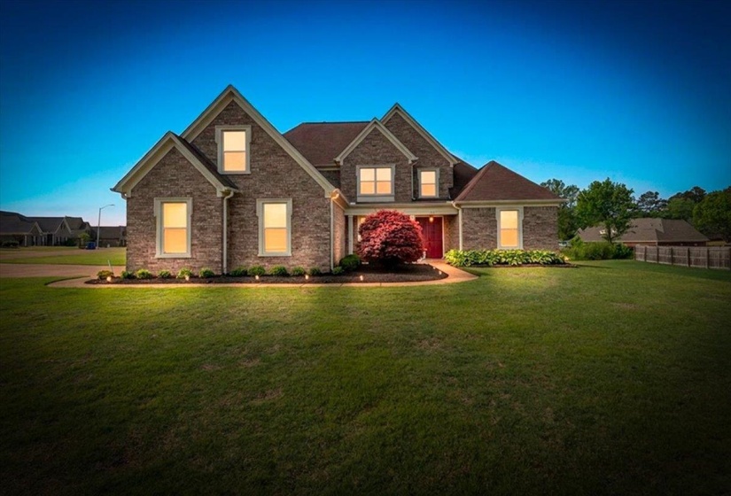 Traditional brick exterior featuring multiple gables, a red front door, accent landscaping, and a manicured lawn with ground lighting