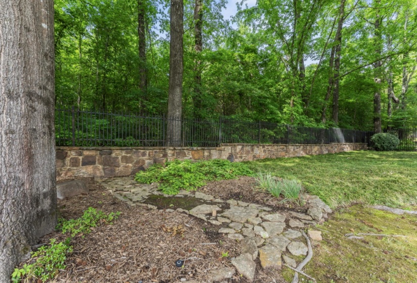 Stone retaining wall topped with a black metal fence, surrounded by mature trees and lush greenery