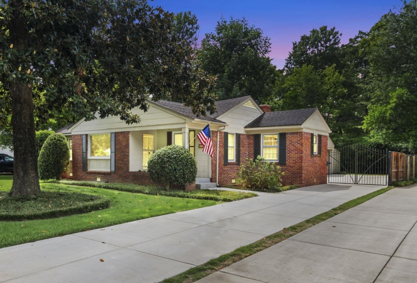 Brick facade residence featuring a front lawn with mature trees and shaped shrubs, a concrete driveway, and a black wrought iron gate