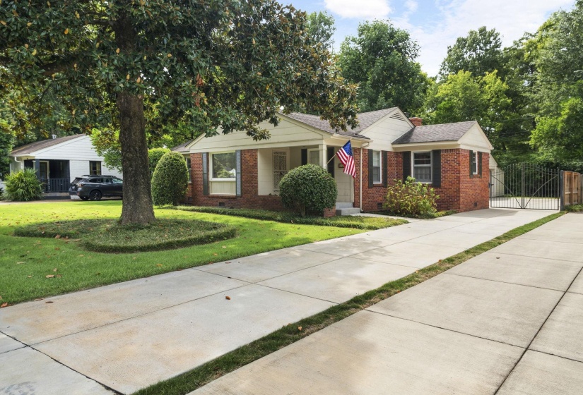 Brick facade residence with expansive concrete driveway, mature front yard tree, and an ornate wrought iron gate
