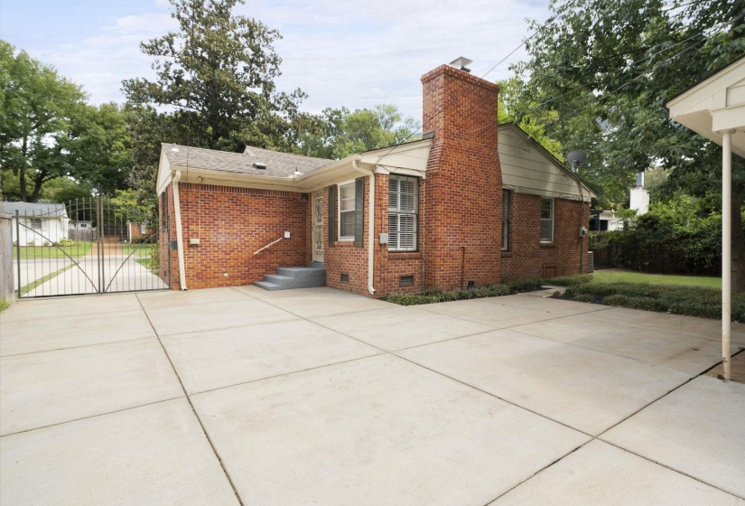 Brick-clad exterior featuring a concrete driveway, a wrought iron gate, a prominent brick chimney, and mature trees providing natural shading
