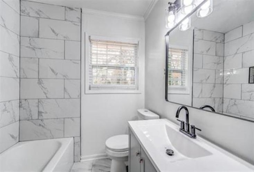 Bathroom featuring a built-in tub with tile surround, a window with blinds, a vanity with an integrated sink, a dark-finish faucet, and a large wall-mounted mirror