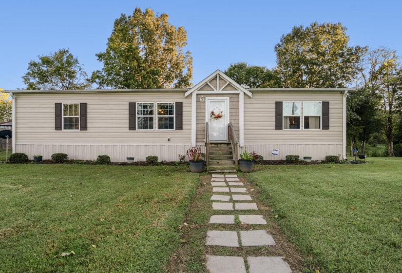 Single-story home featuring light-colored siding, dark shutters, a front porch with natural wood railing, a stone-paver walkway, and surrounding greenery