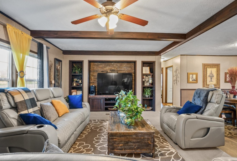 Spacious living area featuring a wood-finish beamed ceiling, a ceiling fan with integrated lighting, and a stone-accented media wall with built-in shelving
