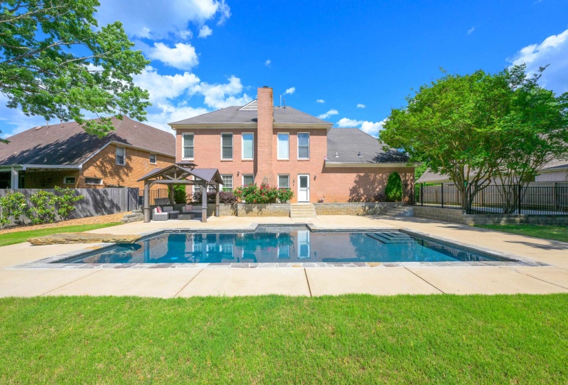 Expansive backyard featuring a rectangular swimming pool, concrete patio surround, a shaded gazebo, and a brick exterior home with a white rear door