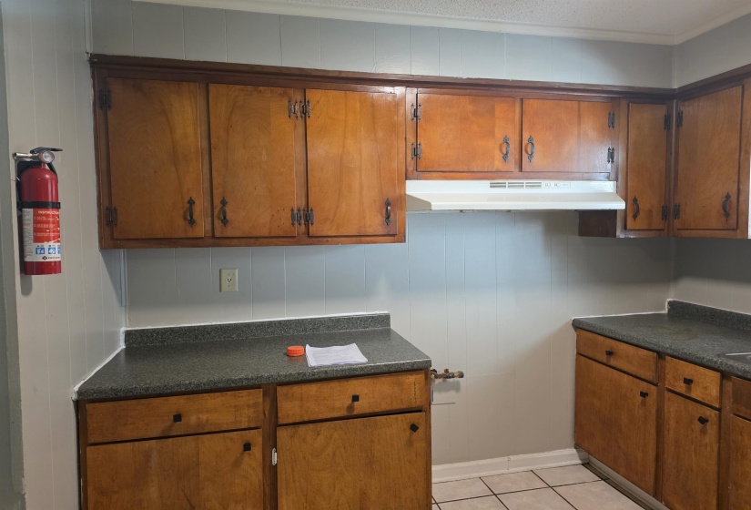 Kitchen with brown cabinetry, dark countertops, light tile patterned floors, under cabinet range hood, and crown molding