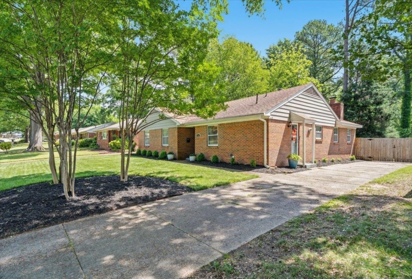 Brick single-story residence featuring a gabled roof, dedicated driveway, mature trees, and a wooden fence