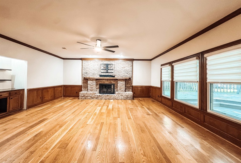 Spacious room featuring wood-finish flooring, a brick fireplace with a wood mantle, wainscoting, and large windows with blinds