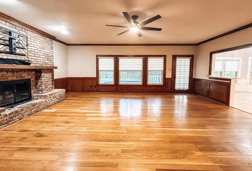 Spacious living area featuring warm wood-finish flooring, a stone-clad fireplace with a hearth and mantel, dark wood wainscoting, and a ceiling fan