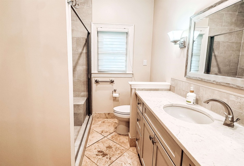 Bathroom featuring a glass-enclosed shower with a built-in bench, a single vanity with an integrated sink and light-colored countertop, decorative wall sconces, a framed mirror, and tile flooring