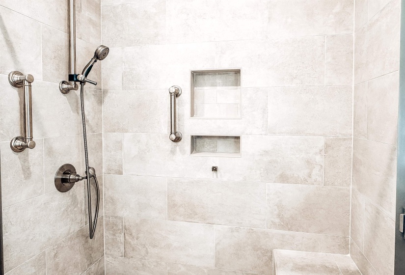Tiled shower enclosure featuring light-toned large format tile, brushed nickel shower fixtures with a handheld spray, integrated wall niches, built-in seating, and grab bars