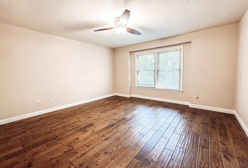 Spacious room featuring wood-finish flooring, neutral-toned walls, a ceiling fan with light fixture, and a large window with blinds