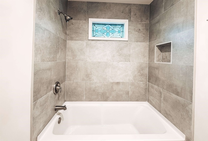 Bathtub with surrounding large format gray tile, oil-rubbed bronze shower fixtures, a decorative stained-glass window, and a recessed shower niche