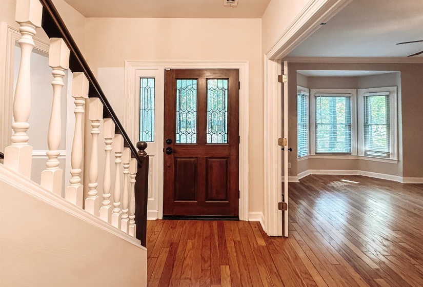 Inviting entryway featuring wood-finish flooring, a solid wood front door with leaded glass sidelights, a classic baluster staircase, and a doorway opening to a room with a bay window