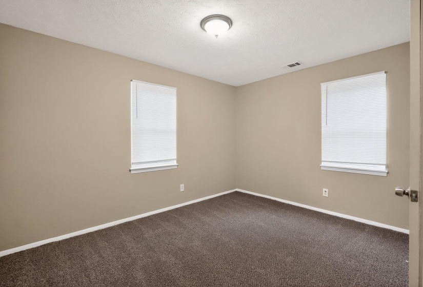 Neutral-toned room featuring a textured carpet, two windows with white blinds, and a flush-mount ceiling light fixture