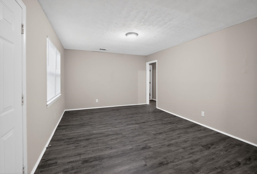 Spacious room featuring dark wood-finish flooring, neutral-toned walls, a single window with blinds, a flush-mount ceiling light fixture, and white trim