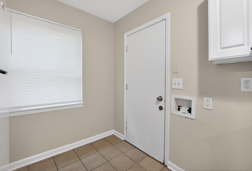 Dedicated utility area featuring a window with white horizontal blinds, a white door, light tan wall paint, and square tile flooring
