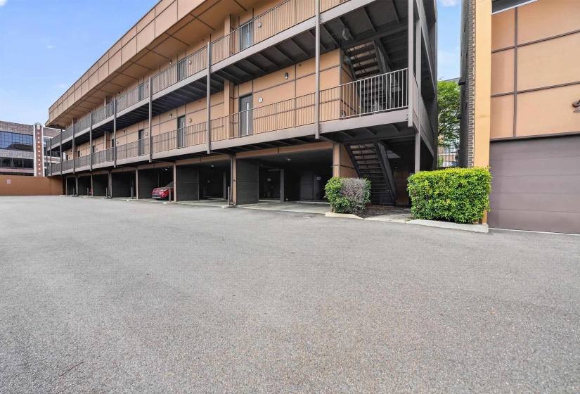 Multi-story building featuring an asphalt parking area, covered parking spaces, individual balconies with dark railings, exterior staircases, and ground-level landscaping