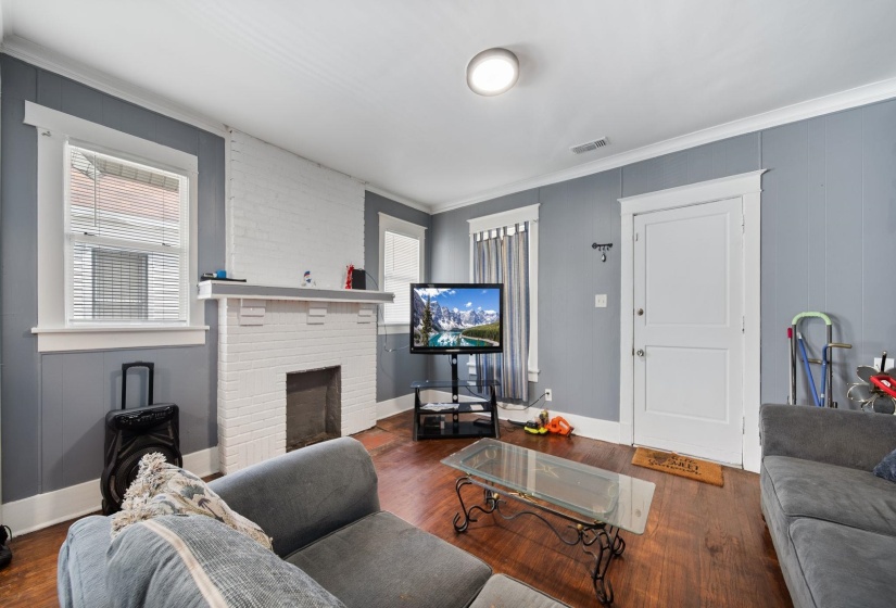 Living area featuring wood-finish flooring, a white-painted brick fireplace with mantel, gray wall paneling, and white trim