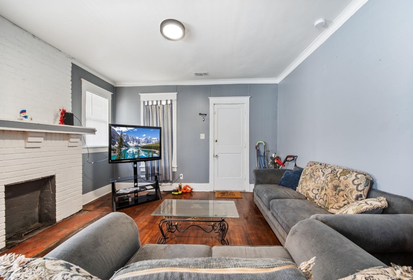 Living area featuring wood-finish flooring, a white brick fireplace, multiple windows with white trim, a flush-mount ceiling light, and crown molding