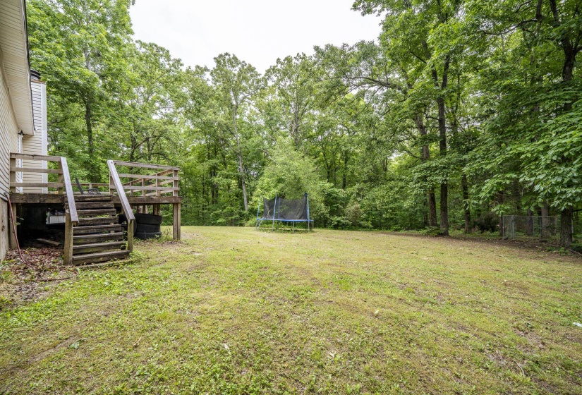 Expansive rear yard featuring a wood-tone deck with stairs and railing, a broad lawn, and mature trees providing natural screening